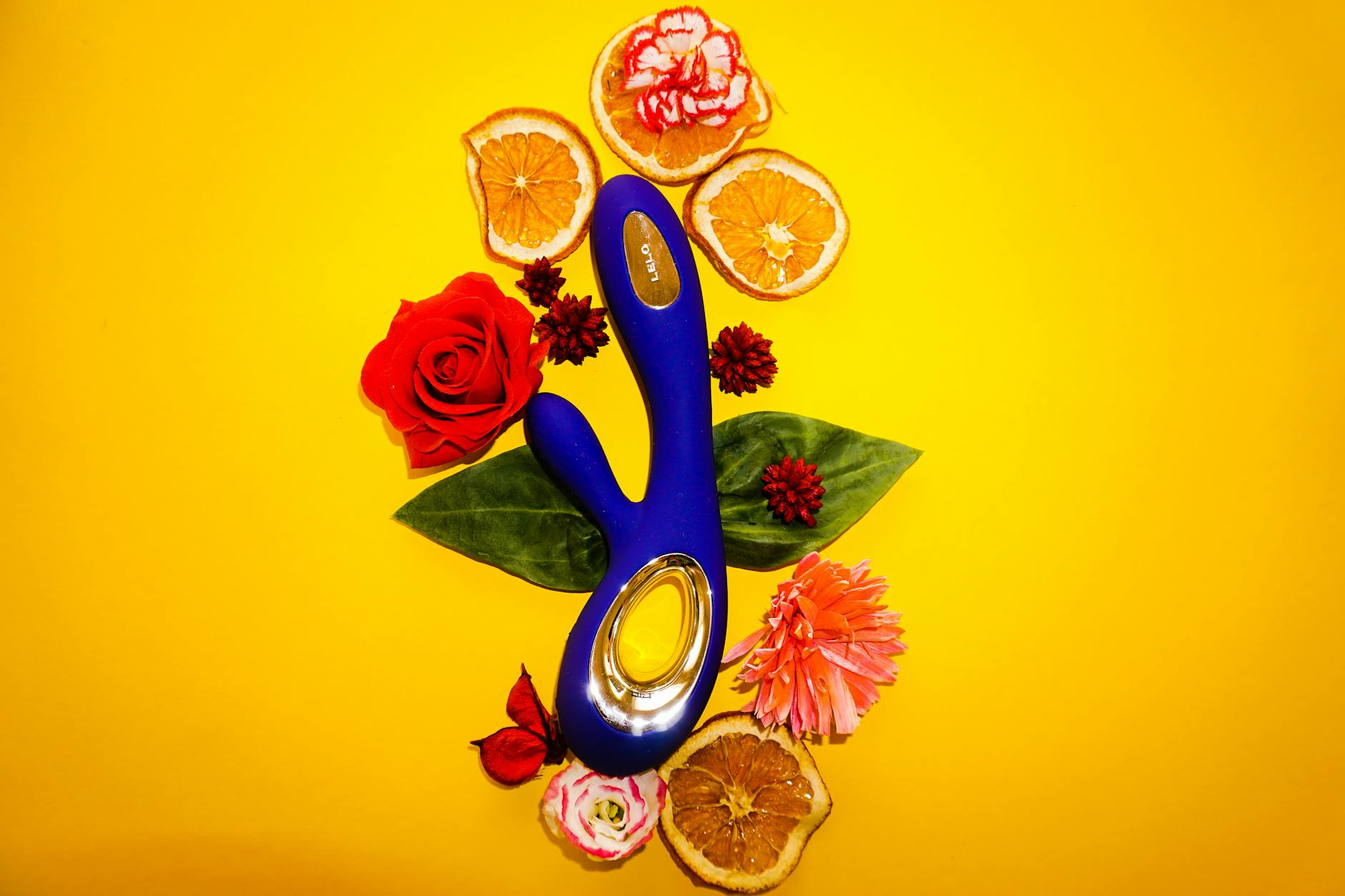 Blue vibrator surrounded by fresh flowers and citrus on a cheerful yellow background